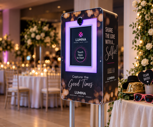 A Lumina wedding event photo booth pod in a decorated event hall, with tables, floral arrangements, and warm string lights in the background. The pod features a black front panel with a glowing purple photo frame, white and gold text, and a side panel with wedding-themed quote graphics.