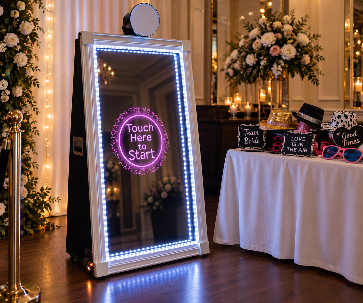 A freestanding illuminated mirror sign with LED border lights and a circular 'Touch Here to Start' graphic, set in a decorated event hall next to a table with novelty hats, sunglasses, and signs